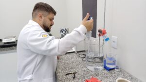 Técnico de laboratório utilizando micropipeta durante ensaio de calibração volumétrica com monitoramento de temperatura em béquer sobre bancada.